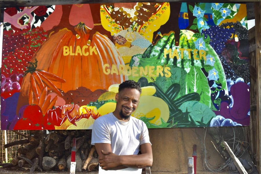 Alumnus Antajuan Adams stands smiling in front of a mural painted at New Roots Urban Farm. 