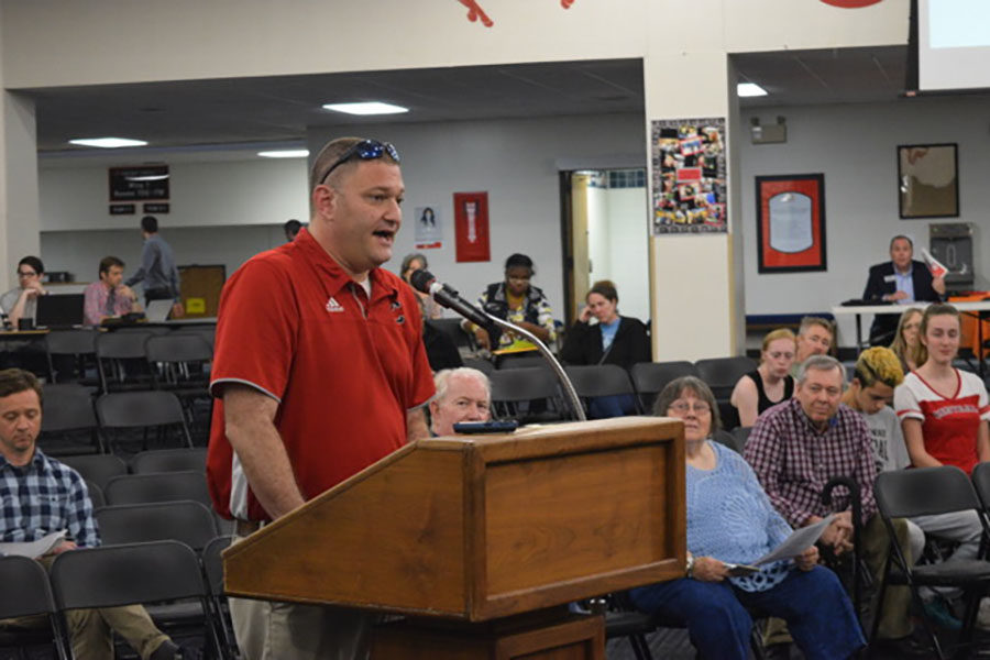 Ken Susman stands in front of the board of education in the foyer of Central Middle school. He is wearing a red shirt and speaking into the microphone at the podium, empassioned.