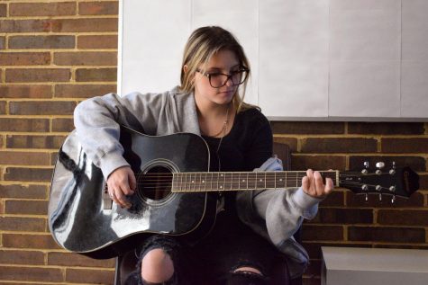 Playing an acoustic guitar, freshman Noelle McSpadden sits just outside of her music classroom. McSpadden was inspired to play guitar to add to her songwriting. “[Guitar class] makes songwriting much easier for me. I feel good about my music because I can actually play with a guitar [to back it up],” McSpadden said.