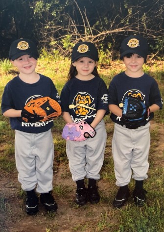The young-triplets-now-seniors Kennedy, Drew and Cole Whitaker, stand side by side holding their baseball gloves and wearing their uniforms. 