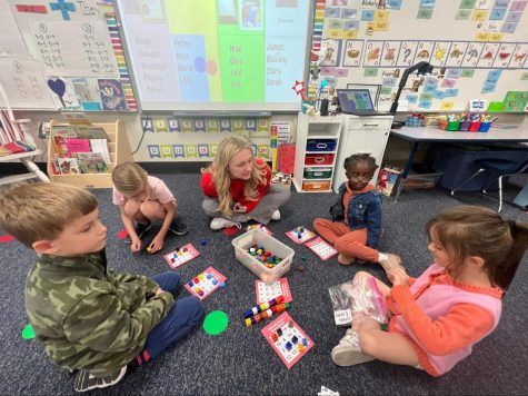 Senior Kelsea Wilson sits on the floor of a kindergarten classroom with four kindergarteners around her. They are playing bingo using colorful place value blocks.