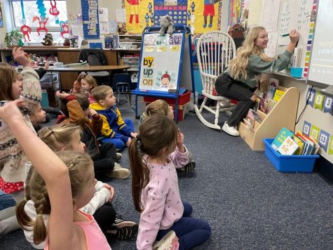 Senior Kelsea Wilson sits at the front of a room on a rocking chair, writing on the white board. A group of kindergarteners watch her, raising their hands to participate.