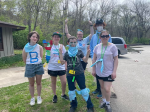 A group of Camp Rainbow attendees stand together outside, wearing colorful t-shirts and bandanas.