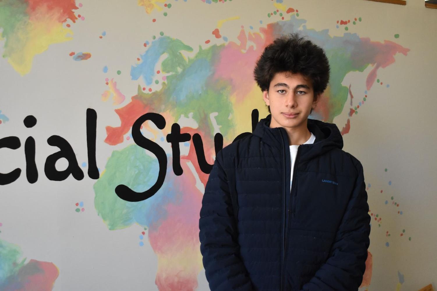 Junior Nicholas Herman stands in front of a white wall with a colorful mural of a world map painted on it. Herman stands in front of the Eastern Hemisphere.