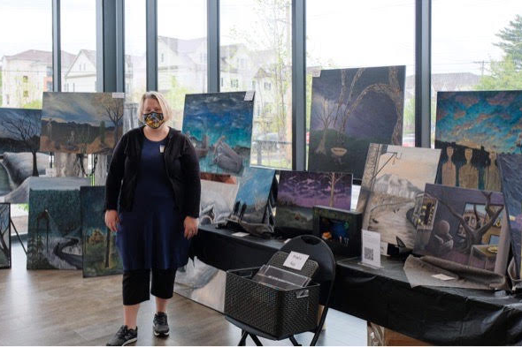 Design Arts and Digital Design teacher Kristi Ponder stands in a room surrounded by many paintings that she has done. The room is large with lots of windows.