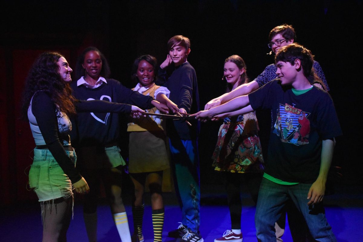 Seven+cast+members+stand+in+a+semi-circle+on+stage%2C+placing+their+wands+in+the+middle+of+their+huddle.