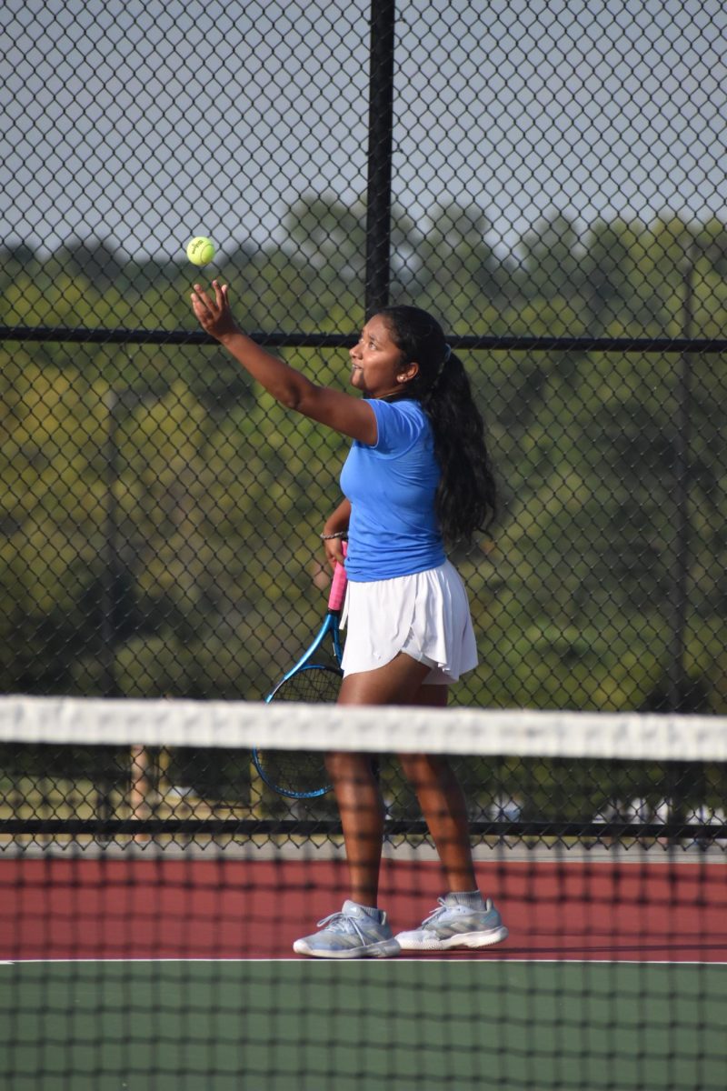 Freshman Mireya David tosses a tennis ball upward, staring at it, with her racket in her other hand. She stands in the center of the court, showing her intense focus.