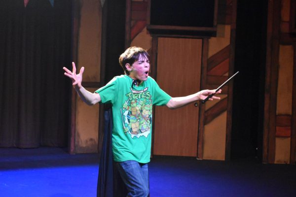 Wearing a green Ninja Turtles shirt, freshman Tate McKenna puts on a sour face with his arms outstretched while on stage, holding a wand. 