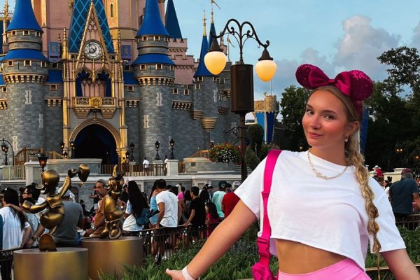 Junior Karry Nickel stands to the right of the photo with her arms outstretched, displaying her pink Minnie Mouse ears. In the background, the Disney Castle stands with a crowd around it.