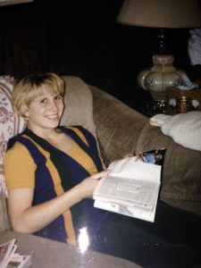 Science teacher Chloe Gallaher smiles at the camera from her couch, holding a book open in her lap.