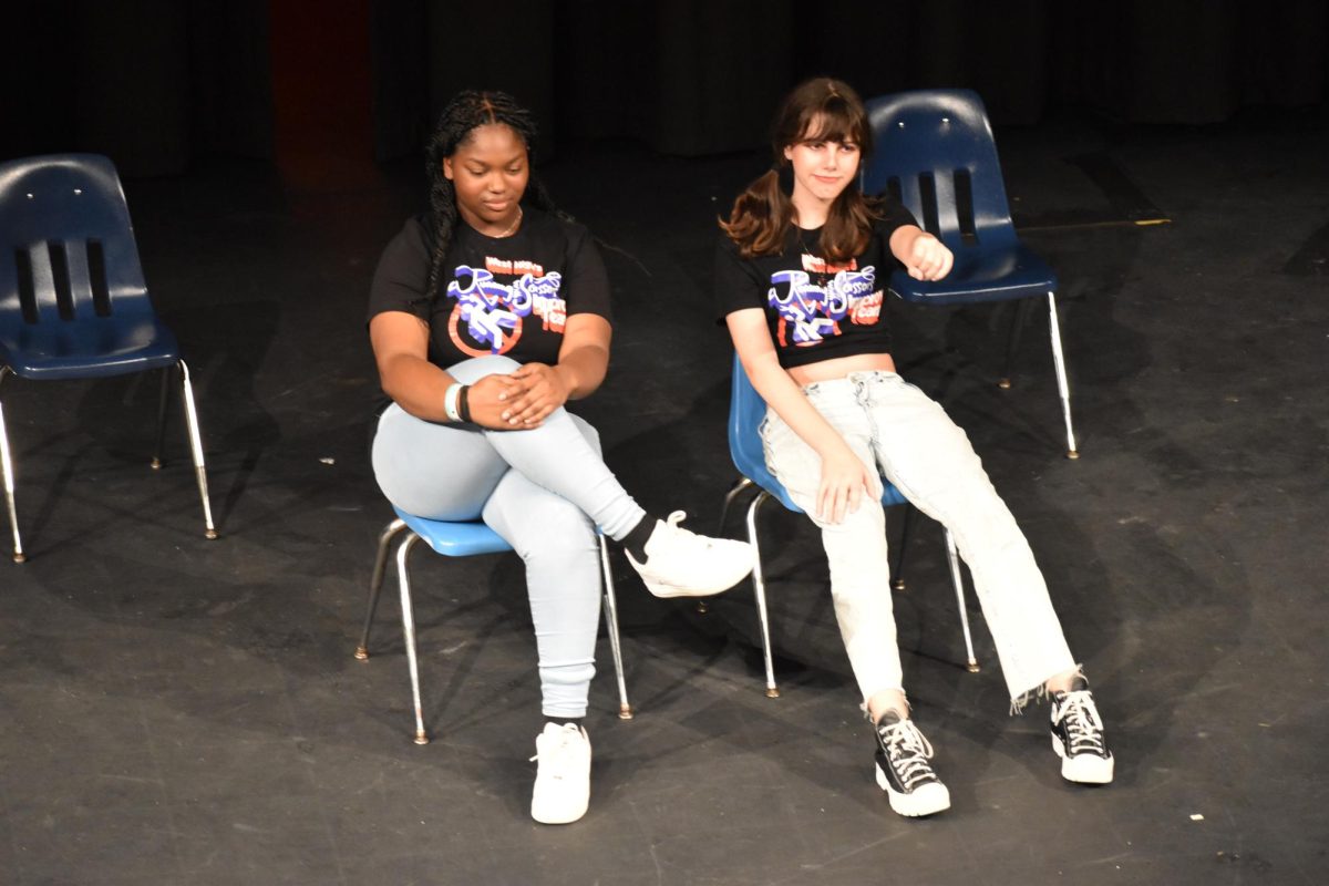 Sophomore Kanya Livingston and junior Ash Herring sit on chairs on stage. Herring extends an arm, pretending to drive a car.