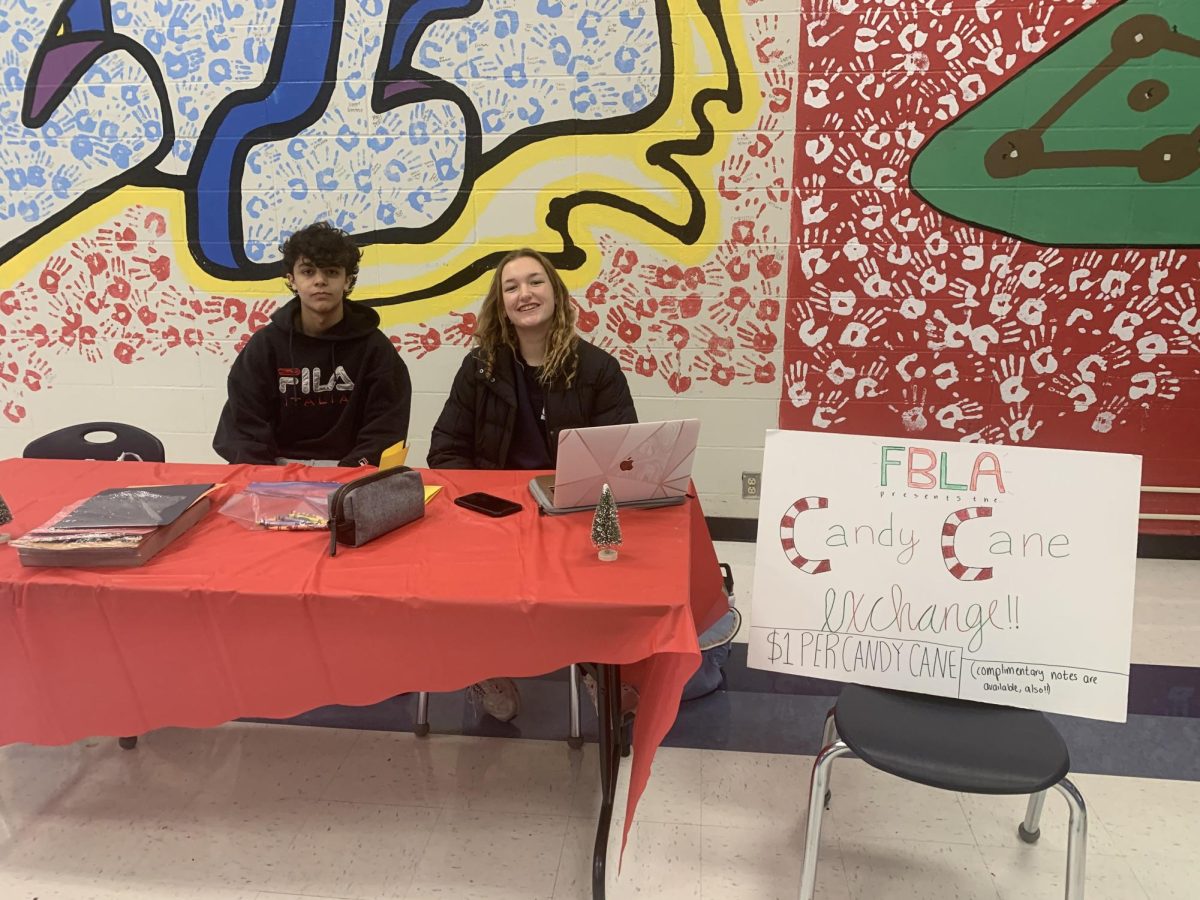 Juniors Laith Alhamid and Brooke Hoenecke smile at the camera, sitting behind a red table in the cafeteria as they sell candy canes. A sign beside them advertises the Candy Cane Exchange.