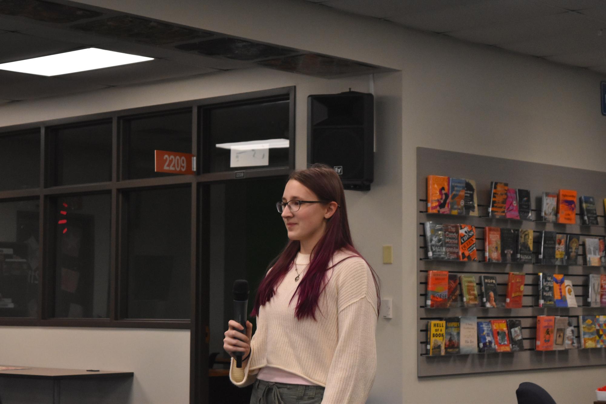 Sophomore Victoria Gold holds a microphone and stands in the library, looking out towards the judges as she faces the crowd.