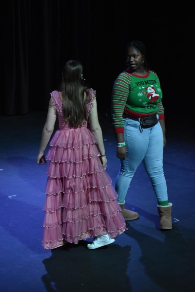 Sophomores Kanya Livingston and Juliana Rodgers look at each other as they stand on stage. Rodgers dons a pink dress while Livingston wears a Christmas sweater.