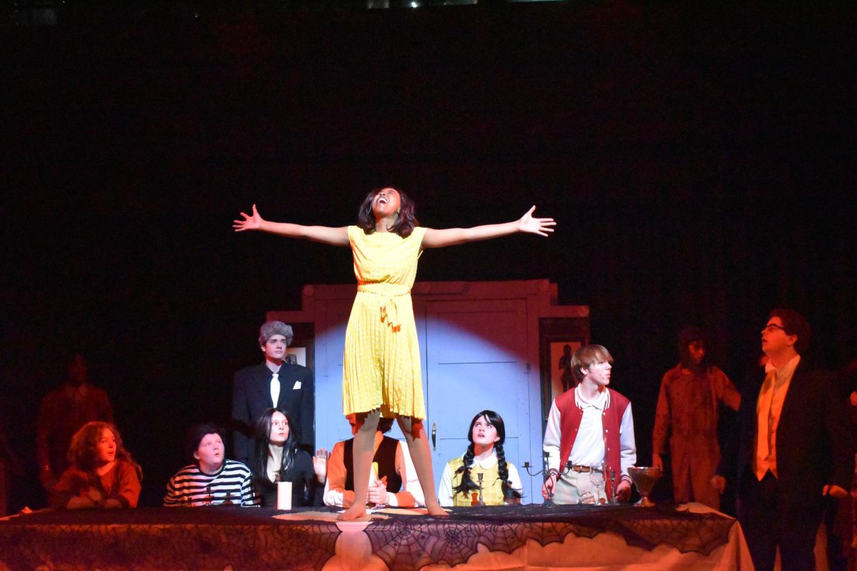 Under the spotlight, senior Mya Jenkins sings “Waiting” on top of the dinner table. Jenkins was most supported by seniors Esta Kamau and Jack Mullen throughout this experience. “Theatre is [a] community. It’s a community of weirdos that come in all different shapes and sizes, but when they are all put together on stage; it’s genuinely so magical to watch,” Jenkins said. 
