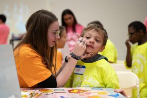 Carefully holding a young athlete's face, senior Elizabeth Knott works at the face painting station in Victory Village. The Victory Village, located in the North Gym, served as a space for athletes to engage in multiple independent tasks. “I like to volunteer [and]  interact with all different kinds of people [and] helping them have a fun experience. There was a little boy who asked me if I could [draw] a character from a game he liked [and] I was able to look it up and draw that character for him,” Knott said. 